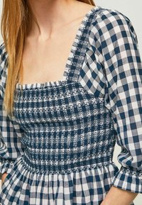 Abito in gingham blu e bianco con corpino smock, scollatura quadrata e maniche a palloncino. Presenta gonna arricciata e cuciture testurizzate.