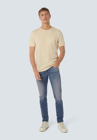 Lichtbeige korte mouwen t-shirt met borstzak, gecombineerd met vervaagde blauwe spijkerbroek en witte sneakers, staand tegen een lichte achtergrond.