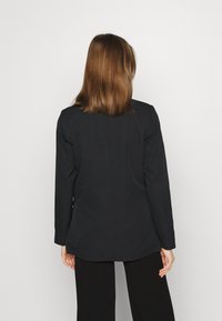 Blazer noir à manches longues avec une coupe ajustée et une texture lisse, présentant un ourlet légèrement incurvé et des détails de couture subtils à l'arrière.