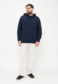 Felpa blu navy con cappuccio e lacci, tasca frontale e dettaglio del logo. Indossata con pantaloni di colore chiaro e sneakers nere. Texture liscia.