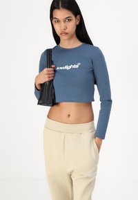 Avkutt blå langermet topp med hvit logo, kombinert med beige joggebukser med ribbestrikket midjebånd og sidelommer. Svart håndveske inkludert.