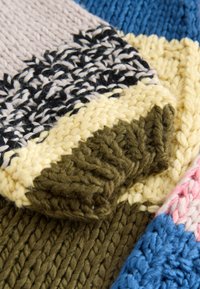 Knit textile featuring a patchwork of colors: beige, black, yellow, green, and blue. Thick, textured yarn with varying stitch patterns.