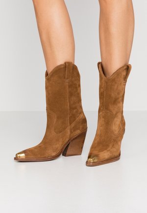 Bottes de cowboy en daim marron avec des talons empilés et des embouts en métal doré portées sur des jambes nues devant un fond léger uni.