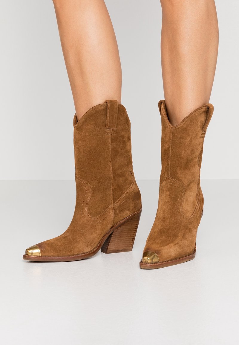 Botas de vaquero de ante marrón con tacones apilados y puntas de metal dorado, usadas sobre piernas desnudas contra un fondo claro.