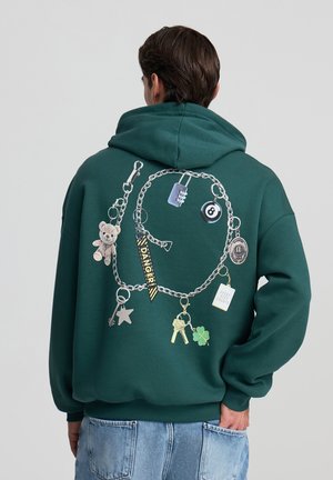 Felpa verde con cappuccio caratterizzata da una grafica a catena decorata con vari charm, tra cui un orsacchiotto, un lucchetto, una stella e un trifoglio, in un design giocoso.
