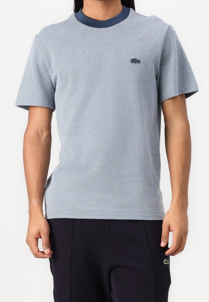 Homme portant une chemise bleu clair à manches courtes avec un col bleu foncé et un pantalon foncé, debout devant un fond uni.