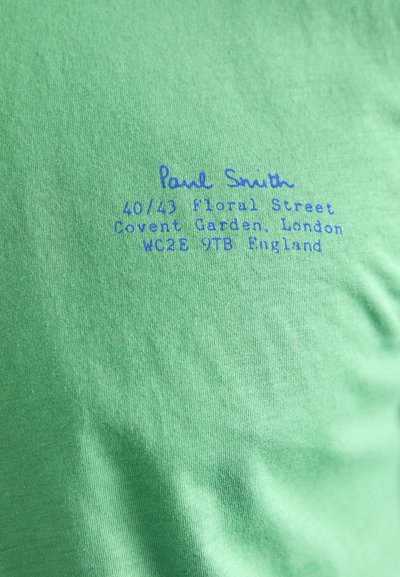 Žalia medvilninė marškinė su mėlynu spaudiniu. Spaudinyje yra "Paul Smith" ir adresas Kavent Gardene, Londone, parašytas neformaliu šriftu.
