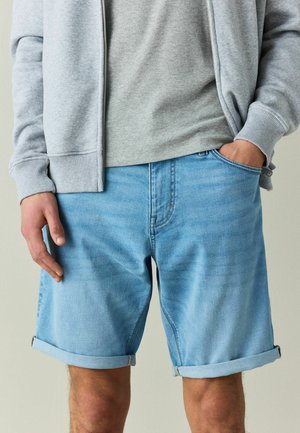 Mann trägt hellblaue hochgekrempelte Jeansshorts und einen grauen Zip-up-Sweatshirt, mit einer Hand in der Tasche und der anderen entspannt.