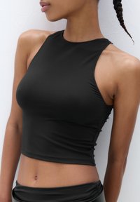Svart kortad tanktop i mjukt tyg, med hög halsringning och halterneckdesign, med en figurnära silhuett som framhäver midjan.