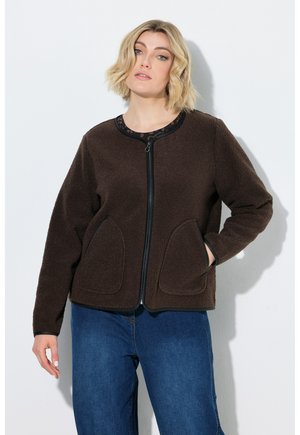 Woman with short blonde hair wearing a dark brown zip-up fleece jacket and blue jeans standing against a white background.