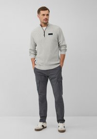 Hellgrauer Pullover mit einem Viertelreißverschluss und einem dunkelblauen Brustpatch, kombiniert mit dunkelgrauen Cargo-Hosen und weißen Sneakern. Glatte Textur, lässige Passform.