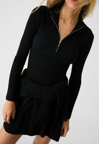 Robe noire côtelée à manches longues avec un col zippé, associée à un pull en maille torsadée vert noué à la taille.