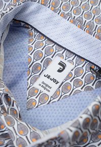 Gekleed shirt met een kraag, gevoerd met een blauw stippenstof. Bevat een tennisracket graphic in grijs en oranje op een witte achtergrond. Label zichtbaar.