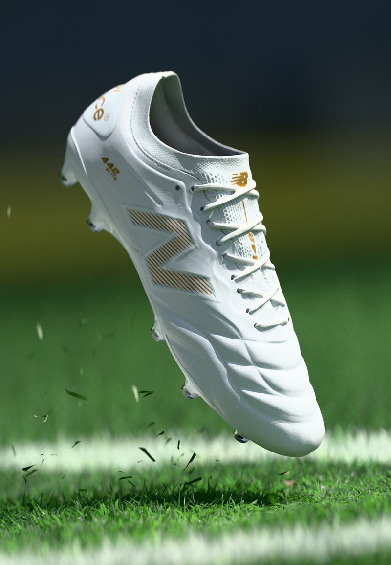 Bijele nogometne kopačke s teksturiranom površinom, opremljene zlatnim akcentima i istaknutim logotipom "N". Lagan dizajn, pogodan za travnate površine.