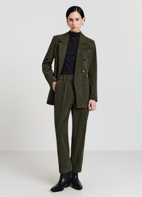 Olivgrüner Cordanzug mit einem doppelt geknöpften Blazer und maßgeschneiderten Hosen, kombiniert mit einem schwarzen Rollkragenpullover und schwarzen Stiefeletten.