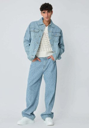 Jeune homme portant une veste et un pantalon en denim bleu clair à motifs coordonnés, un pull en maille blanc et des baskets blanches, debout les mains dans les poches.