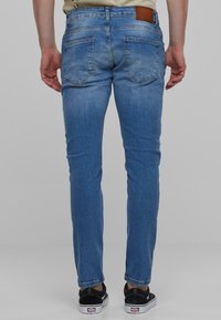 Világoskék, slim fit farmernadrág, hátulsó zsebekkel és barna bőr címkével a derékrésznél. Fekete sportcipővel viselve.