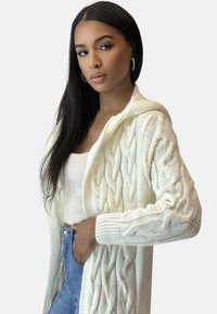 Weißer gestrickter Cardigan mit Kabelmustern, Kapuze, offenem Frontbereich und gerippten Bündchen, kombiniert mit einem hellen Tanktop und Jeans.