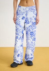 Pantalon à motifs bleu et blanc avec un design floral, coupe décontractée, tissu texturé et poches latérales. Porté avec des chaussures noires.
