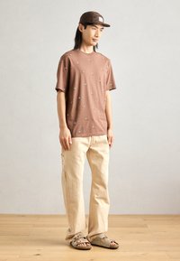 Brun kortærmet T-shirt med små, broderede fugle; parret med beige, løstsiddende bukser og brune sandaler med flere stropper.