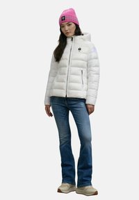 Vrouw in een witte pufferjack, blauwe jeans, witte sneakers en een roze beanie, staand tegen een effen achtergrond.