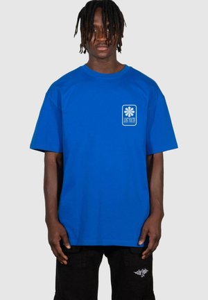 Blå bomullströja med en boxlogotyp som har en blommönster och texten "LOST YOUTH", korta ärmar och avslappnad passform. Bärs med svarta byxor.