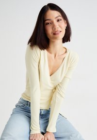 Langærmet, ribbet top i lys gul med dyb V-udskæring og dekorative kanter. Parret med blå denim jeans. Blød tekstur synlig.