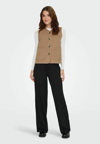 Donna con indosso un gilet color sabbia con bottoni sopra una maglia bianca a maniche lunghe, pantaloni neri a gamba larga e scarpe nere, in piedi davanti a uno sfondo semplice.