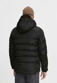 Veste doudoune noire avec capuche, dotée de coutures horizontales et d'une texture douce. Portée sur un pantalon gris, vue de dos.