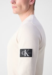Svijetlosmeđa košulja dugih rukava s teksturiranim materijalom, s crnim pravokutnim logotipom na gornjem dijelu rukava koji kaže "Calvin Klein Jeans."