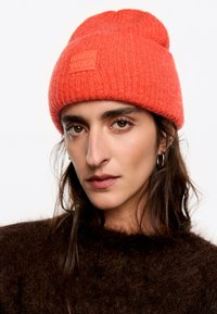Gorro de lana rojo con un patrón texturizado y un parche de goma con marca. Llevado con un suéter oscuro y suave en un retrato en primer plano.