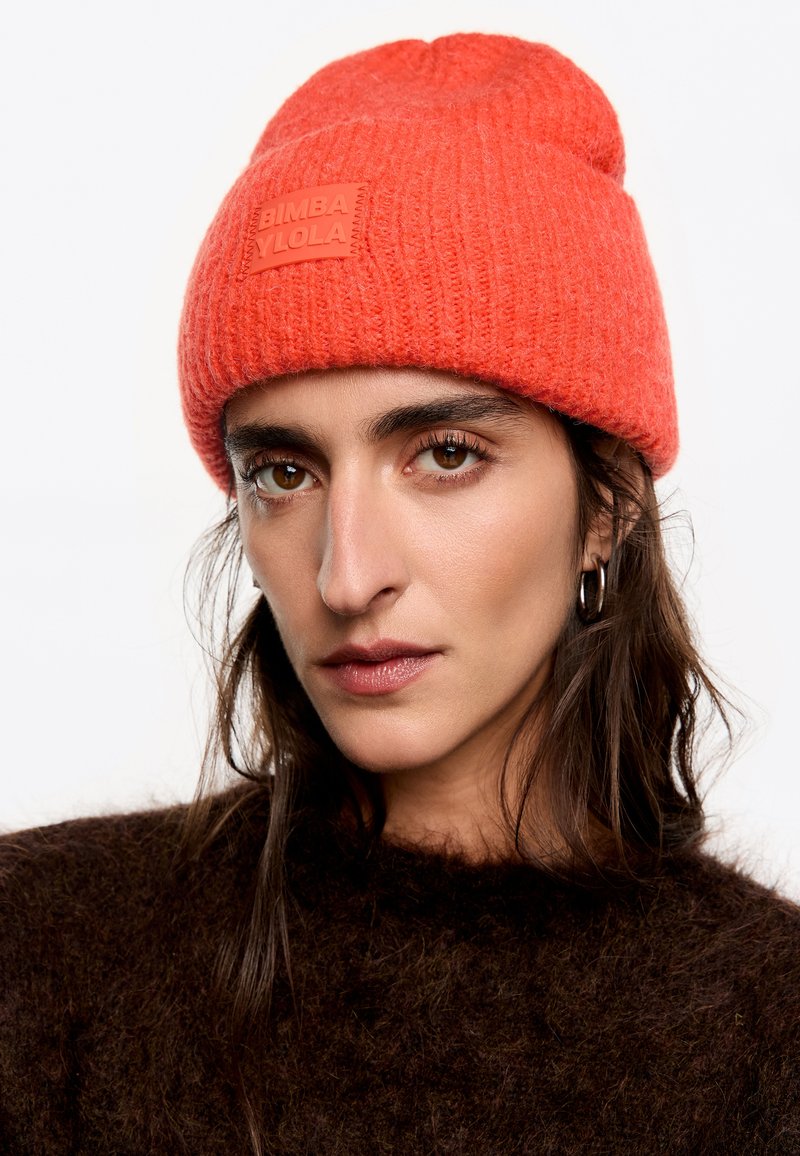 Gorro de lana rojo con un patrón texturizado y un parche de goma con marca. Llevado con un suéter oscuro y suave en un retrato en primer plano.