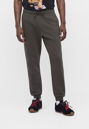Homme portant un pantalon de jogging vert foncé à cordon, des baskets noires avec des lacets rouges, et une partie d'un t-shirt graphique noir.