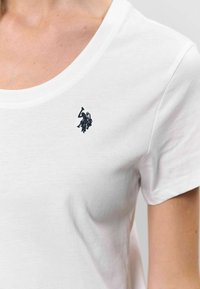 Balts kokvilnas T-krekls ar mīkstu tekstūru, ar mazu tumši zilu izšūtu logo kreisajā krūšu daļā un apaļu apkakli.