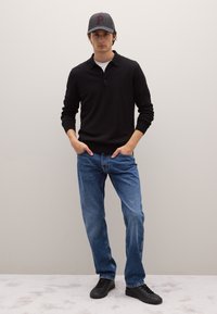 Polo nera a maniche lunghe, jeans blu e sneakers nere. Cappellino grigio con una "P" rossa. Modello in piedi con le mani nelle tasche contro uno sfondo neutro.