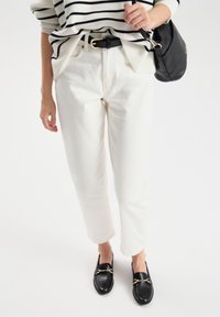 Białe bawełniane spodnie o luźnym kroju, zestawione z paskowanym swetrem z dzianiny i czarnymi mokasynami z złotymi akcentami. W zestawie znajduje się również czarny pasek i torba.