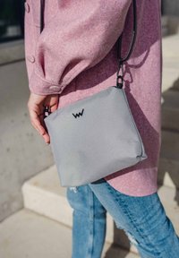 Šedá látková kabelka s texturovaným povrchem, na přední straně s černým logem, kterou drží osoba v růžovém kabátu a modrých džínách.