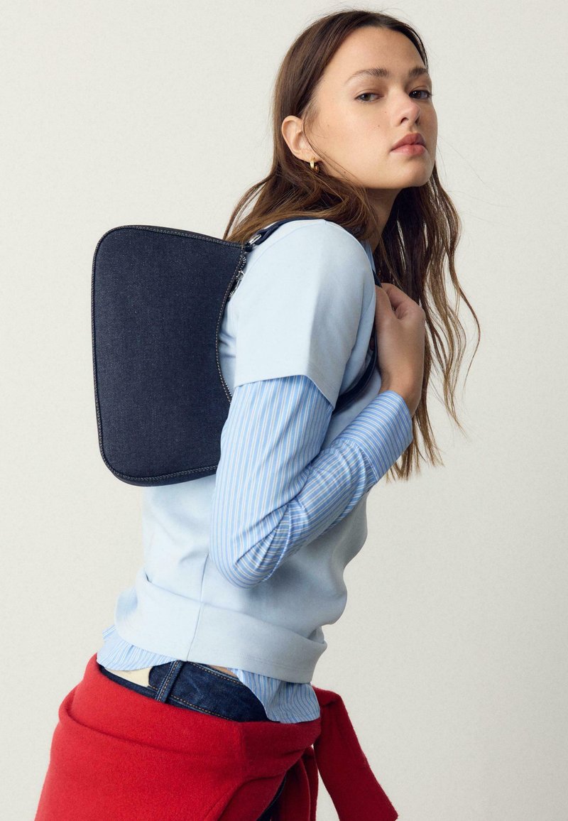 Jeune femme aux longs cheveux portant des chemises bleues superposées et un pull rouge noué autour de la taille, portant un sac à bandoulière rectangulaire bleu foncé.
