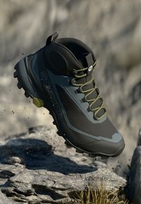 Crne i sive planinarske čizme s teksturiranim gornjim dijelom, zelenim vezicama i robusnom potplatom, uzdignute iznad stjenovitog terena. Sadrže „SCARPA“ oznaku.