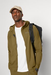 Light olive green zip-up jacket with a hood, paired with a white t-shirt. A black backpack is slung over one shoulder, and a beige cap is worn.