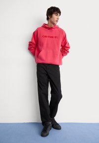 Sudadera rosa con un bolsillo frontal y logotipo bordado; combinada con pantalones negros y zapatillas negras, de pie frente a un fondo neutral.