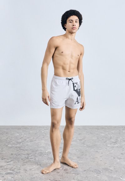 Λευκά μαγιό σορτς με μαύρο κείμενο και γραφικό σχέδιο, ελαστική μέση με κορδόνι, φορεμένα από μοντέλο χωρίς μπλούζα σε ουδέτερο περιβάλλον.