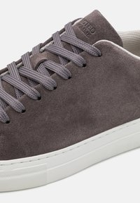 Gray suede sneaker with round white rubber sole, flat gray laces, and subtle branding on the heel. Textured upper and seamless design.