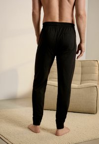 Pantaloni de jogging negri, cu talie elastică, picioare conice și culoare uniformă. Purtați de o persoană care stă pe un covor texturat.