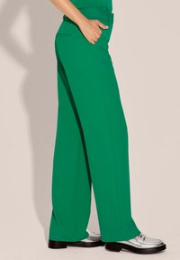 Brede beenbroek in een levendige groene stof, met een hoge taille, zijzakken, strakke lijnen en een zachte textuur. Zilveren schoenen voegen contrast toe.