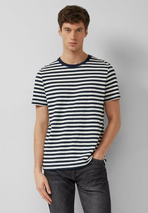 Młody mężczyzna ubrany w granatowo-biały koszulka z krótkim rękawem w poziome paski i ciemne dżinsy, stojący na tle jednolitego jasnoszarego tła.