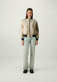 Mladá žena sa usmieva, má na sebe béžovú prešívanú bomber bundu, svetlomodré džínsy a čierne topánky, stojí pred jednoduchým pozadím.