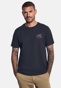 Námořnická modrá bavlněná tričko s bronzovým logem na hrudi a krátkými rukávy; hladká textura, kulatý výstřih, volný střih.