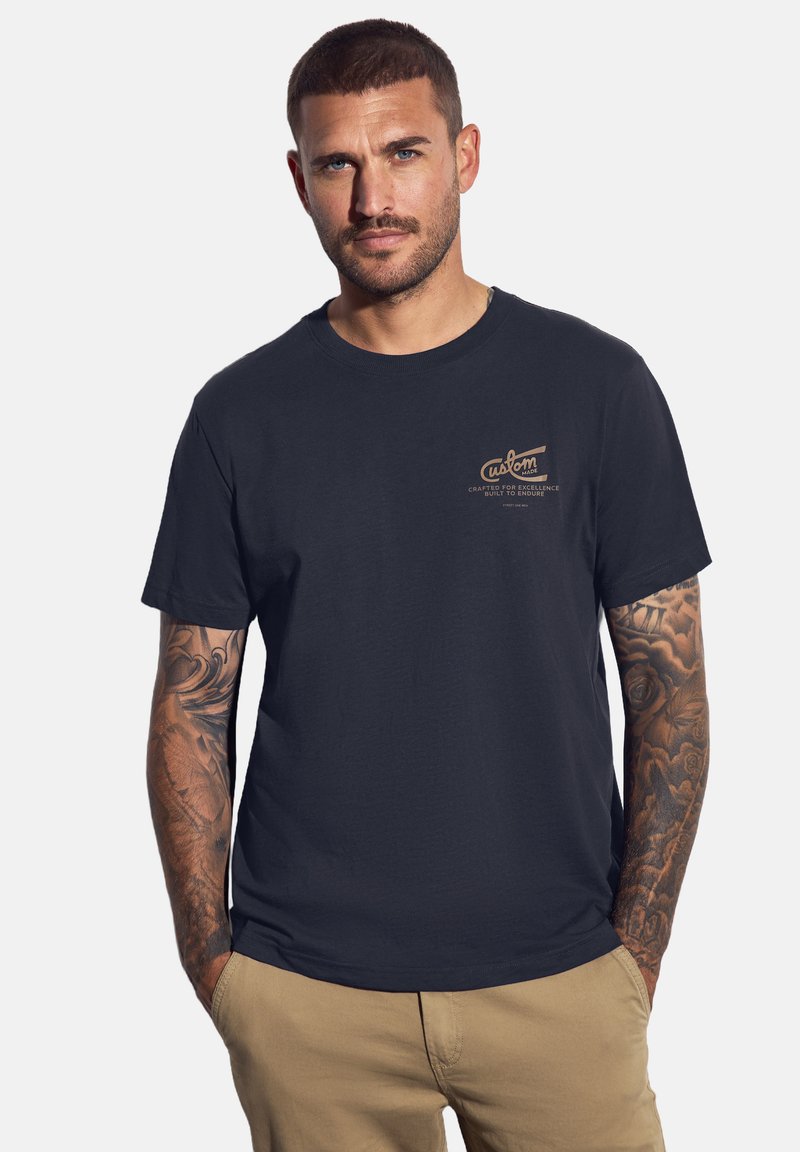 Námořnická modrá bavlněná tričko s bronzovým logem na hrudi a krátkými rukávy; hladká textura, kulatý výstřih, volný střih.