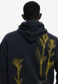 Marineblå hættetrøje med en stor gul blomsterbroderi på ryggen, der har et tekstureret design og en snørehætte.